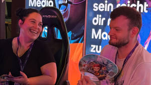 Zwei Teammitglieder der SRH lachen und unterhalten sich am Messestand, im Hintergrund ein Gaming-Stuhl und beleuchtete Werbebanner. Einer der beiden hält einen Fächer mit Spielmotiv in der Hand.